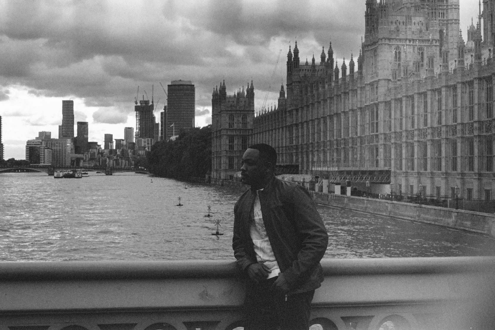 Black and white photograph of a figure at a river embankment rail with the city skyline beyond